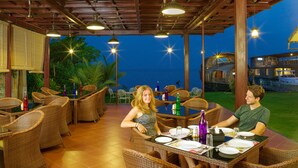 An outdoor dining area with wicker furniture and tables set for two, under a wooden pergola with hanging lights. - Cyrus Resort by Tolins Hotels & Resorts (Cherthala)