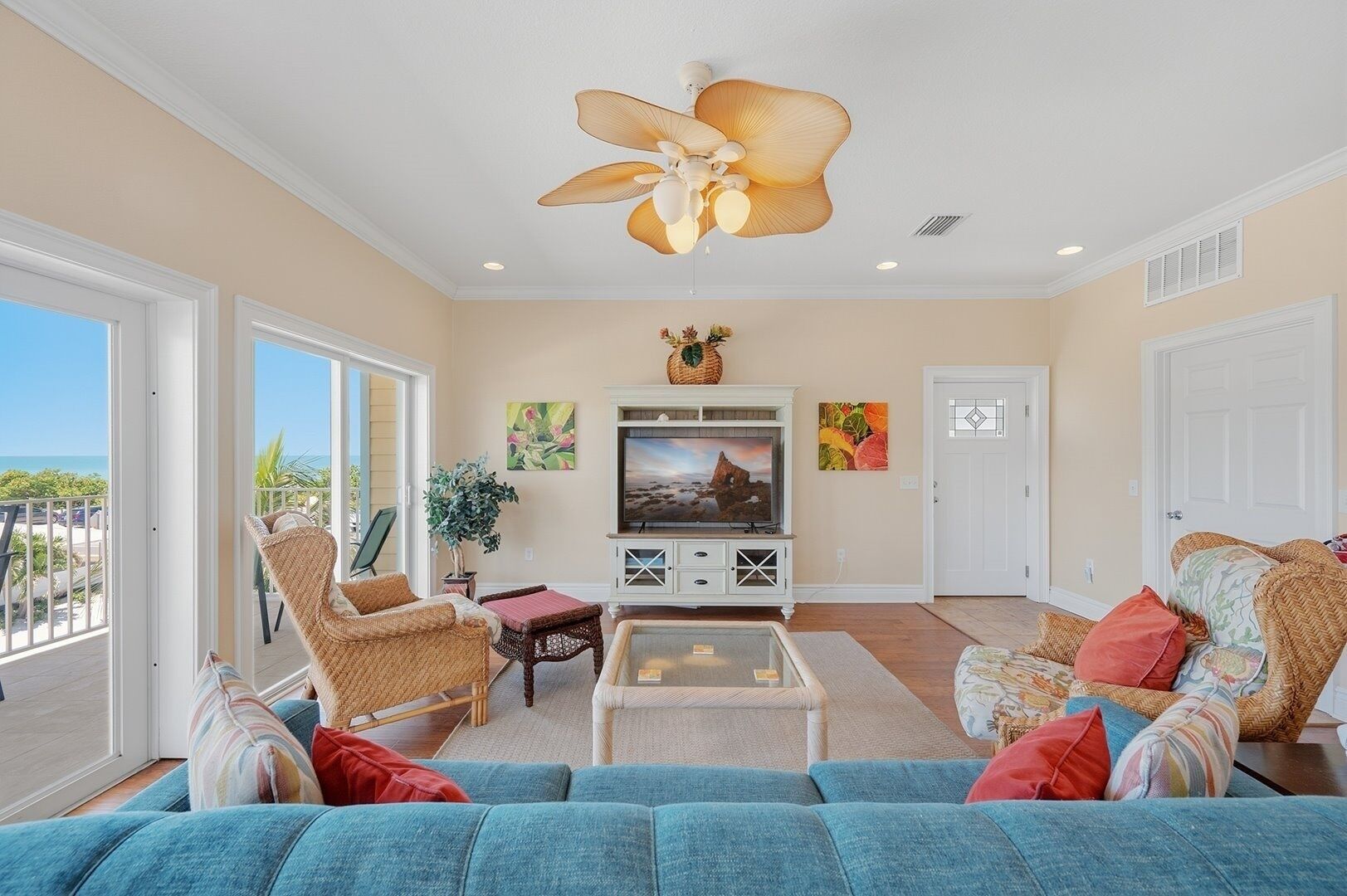 A living room with a large TV, wicker furniture, a ceiling fan, and a view of the outdoors.