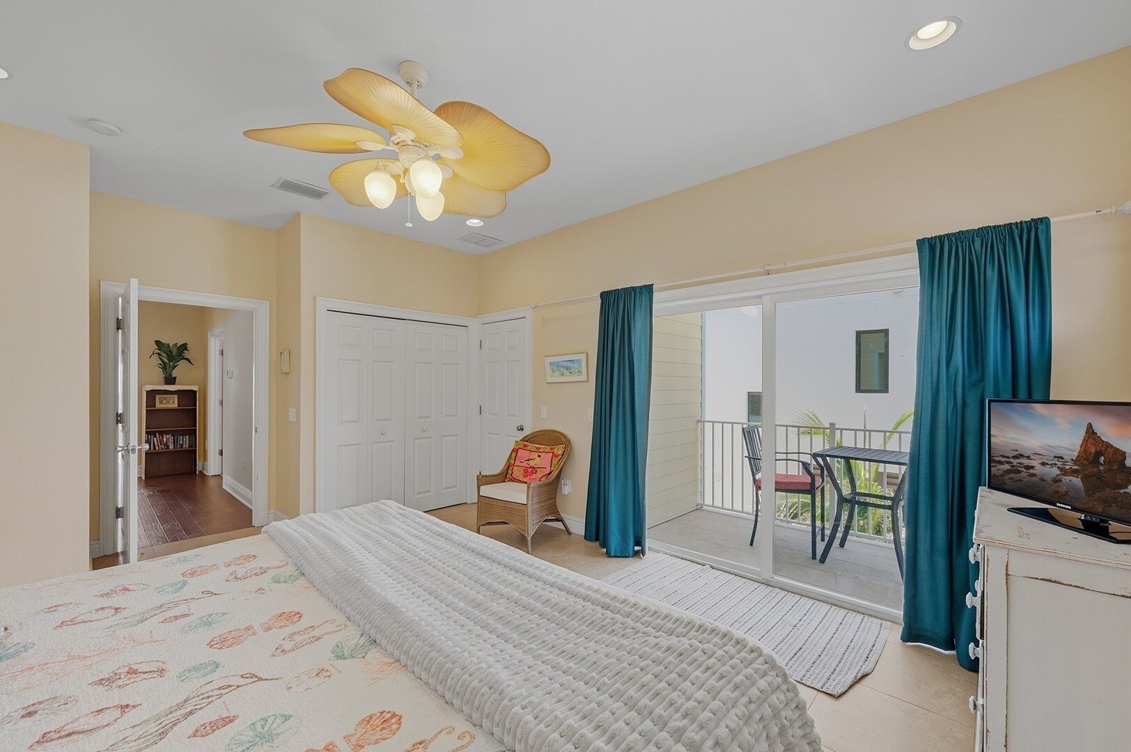 A bedroom with a bed, a ceiling fan, a television, a chair, a balcony with curtains, and a door leading to a hallway.