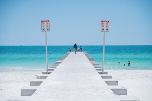 Un muelle que lleva a una playa con agua azul cristalina, gente caminando y dos señales de precaución.