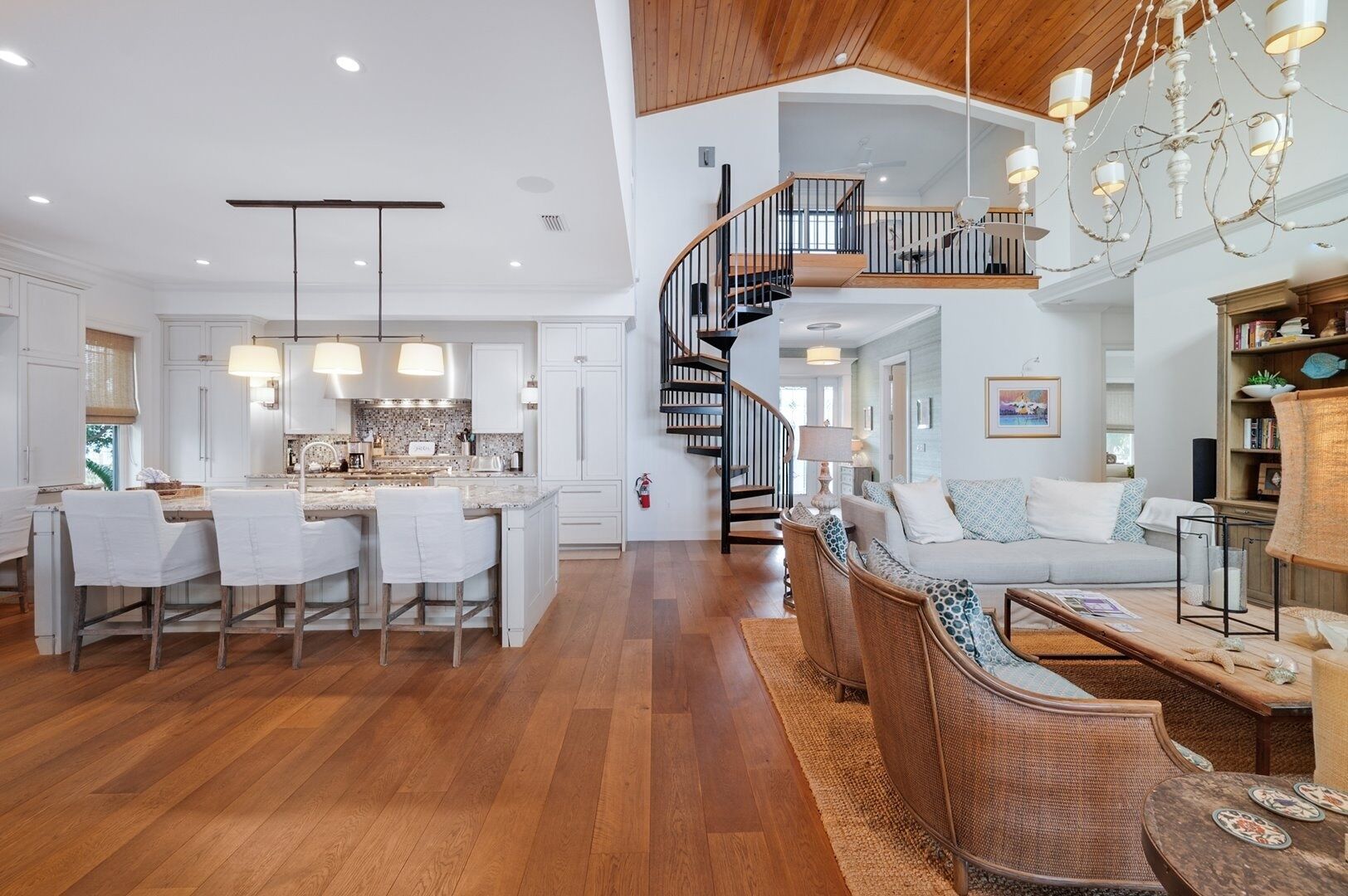 A spacious kitchen with a central island, a dining area with white chairs, a staircase leading to an upper level, and a living room with a sofa and wicker furniture.