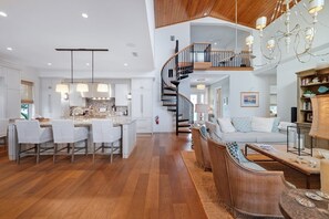 A spacious kitchen with a central island, a dining area with white chairs, a staircase leading to an upper level, and a living room with a sofa and wicker furniture. - Dockside Darling 3 Bedroom Home by RedAwning (Bradenton Beach)