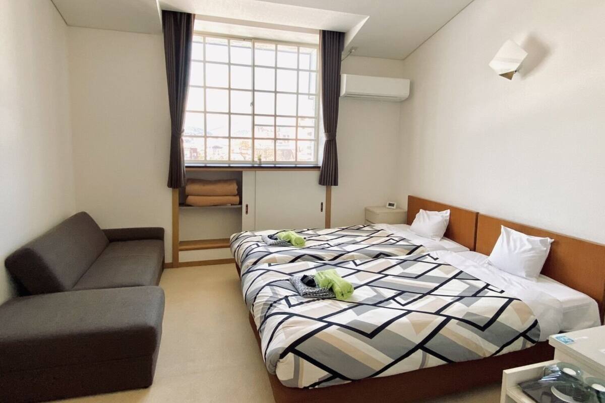 Una habitación de hotel con cama, sofá, ventana con cortinas, aire acondicionado y una pequeña mesa.