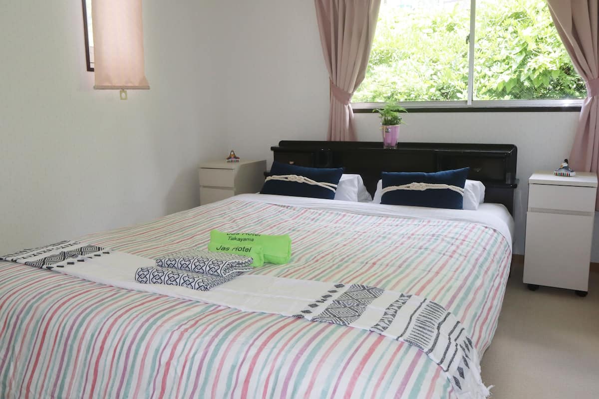Una cama bien hecha con ropa de cama a rayas y almohadas, una mesita de noche con lámpara, y una ventana con cortinas.