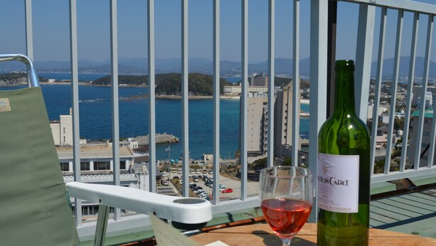 Un balcon donnant sur une ville côtière, une bouteille de vin verte et un verre de vin rouge posés sur une table.