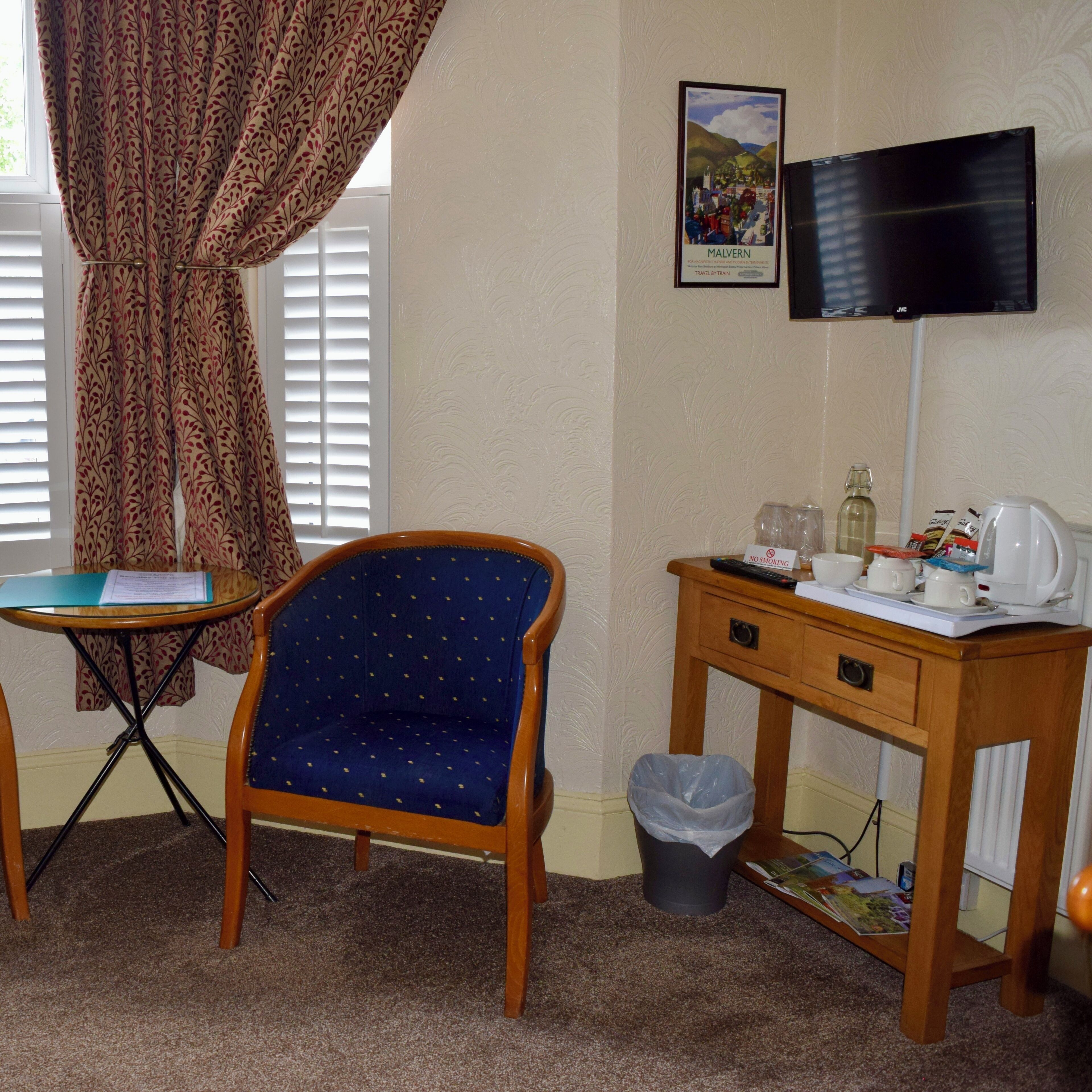 A room with a blue polka dot chair, a small wooden desk with a television, a window with blinds, and a framed picture on the wall.