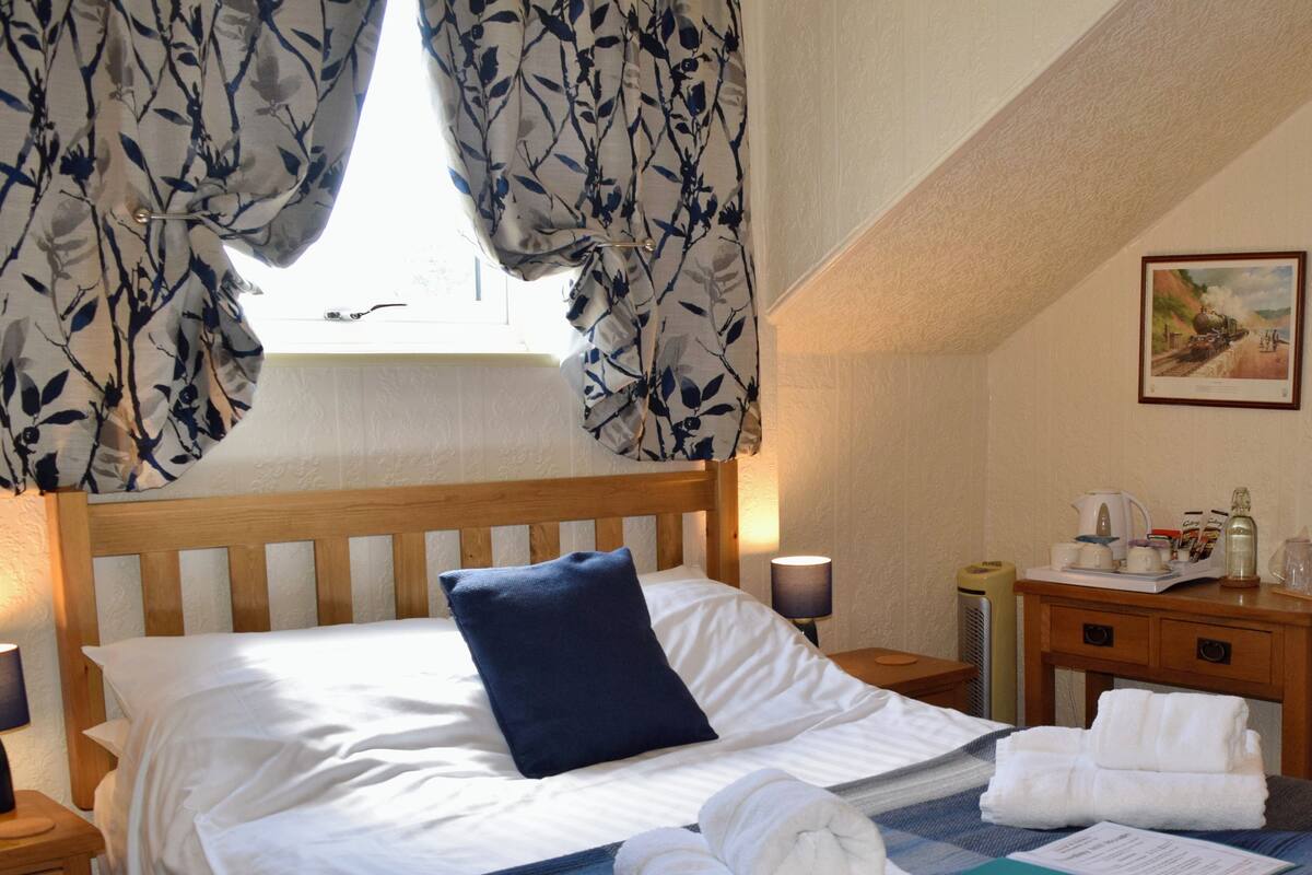 A neatly made bed with white linens, a blue pillow, and a plaid blanket. There are two bedside tables with lamps, a nightstand, and a small wooden dresser with a lamp and a framed picture on the wall.