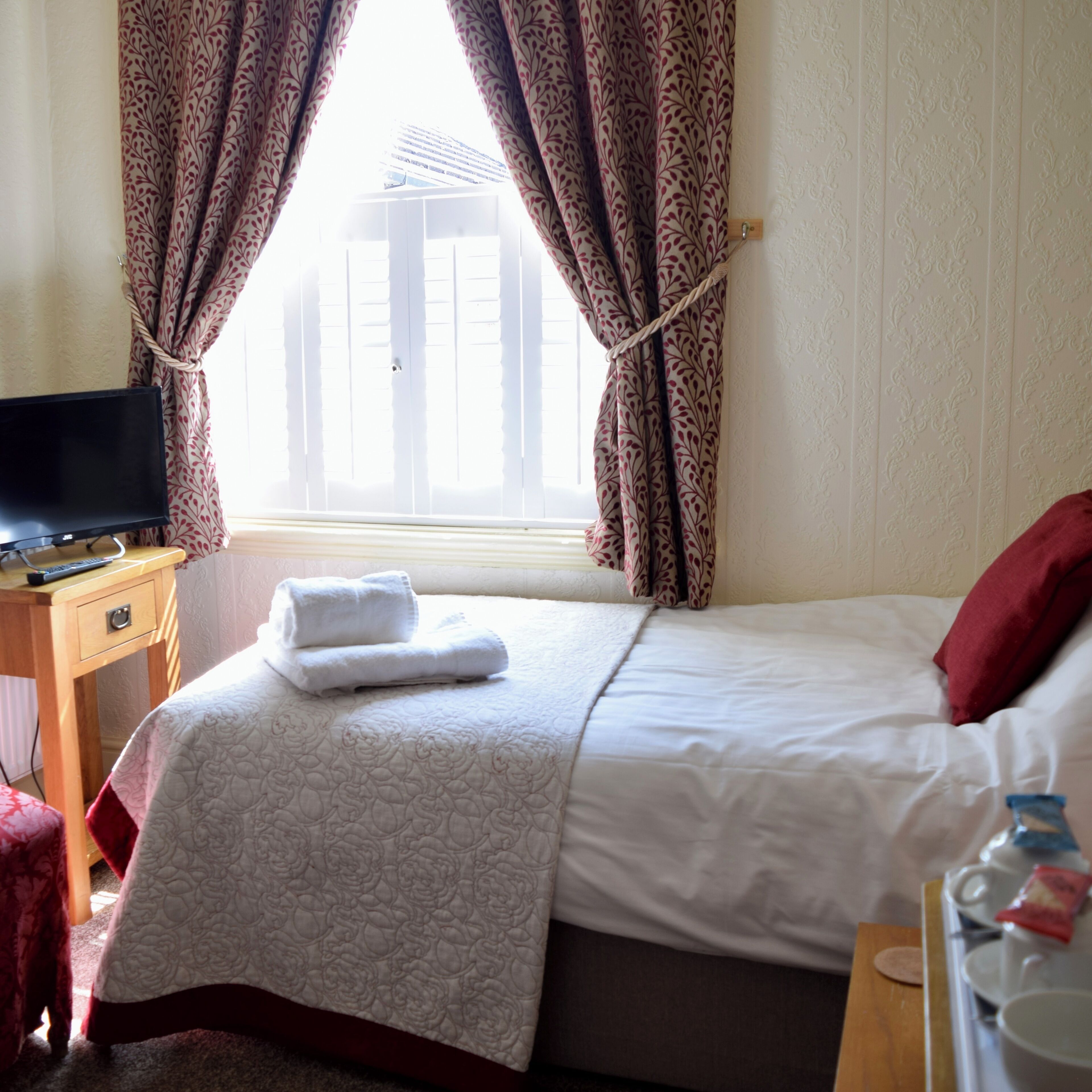 A hotel room with a bed, a TV on a wooden nightstand, a window with curtains, and a bedside table with a tray of breakfast items.