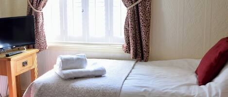 A hotel room with a bed, a TV on a wooden nightstand, a window with curtains, and a bedside table with a tray of breakfast items.