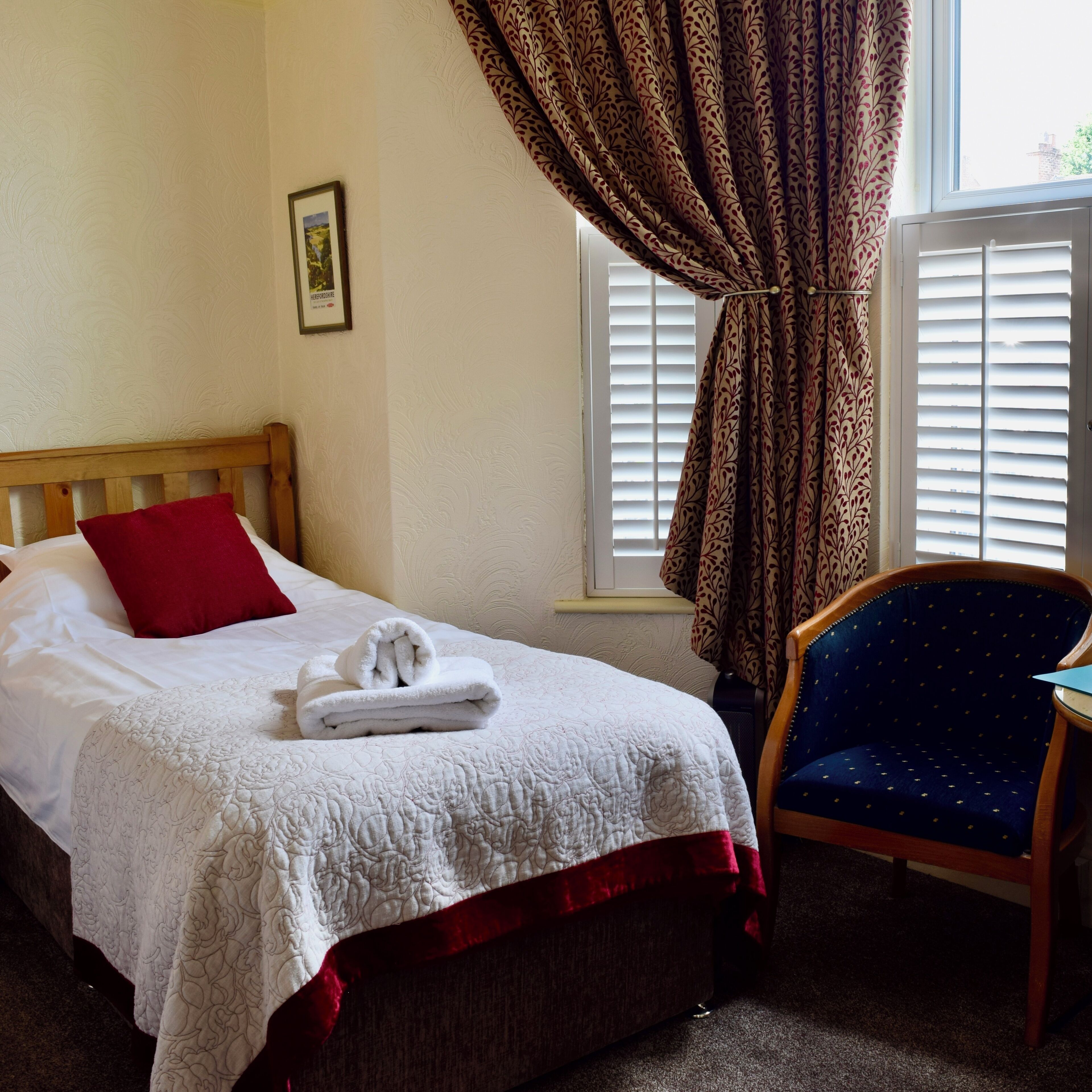 A neatly made bed with a white duvet, a red pillow, and a folded towel. There is a wooden headboard, a blue upholstered chair, and a window with closed blinds.