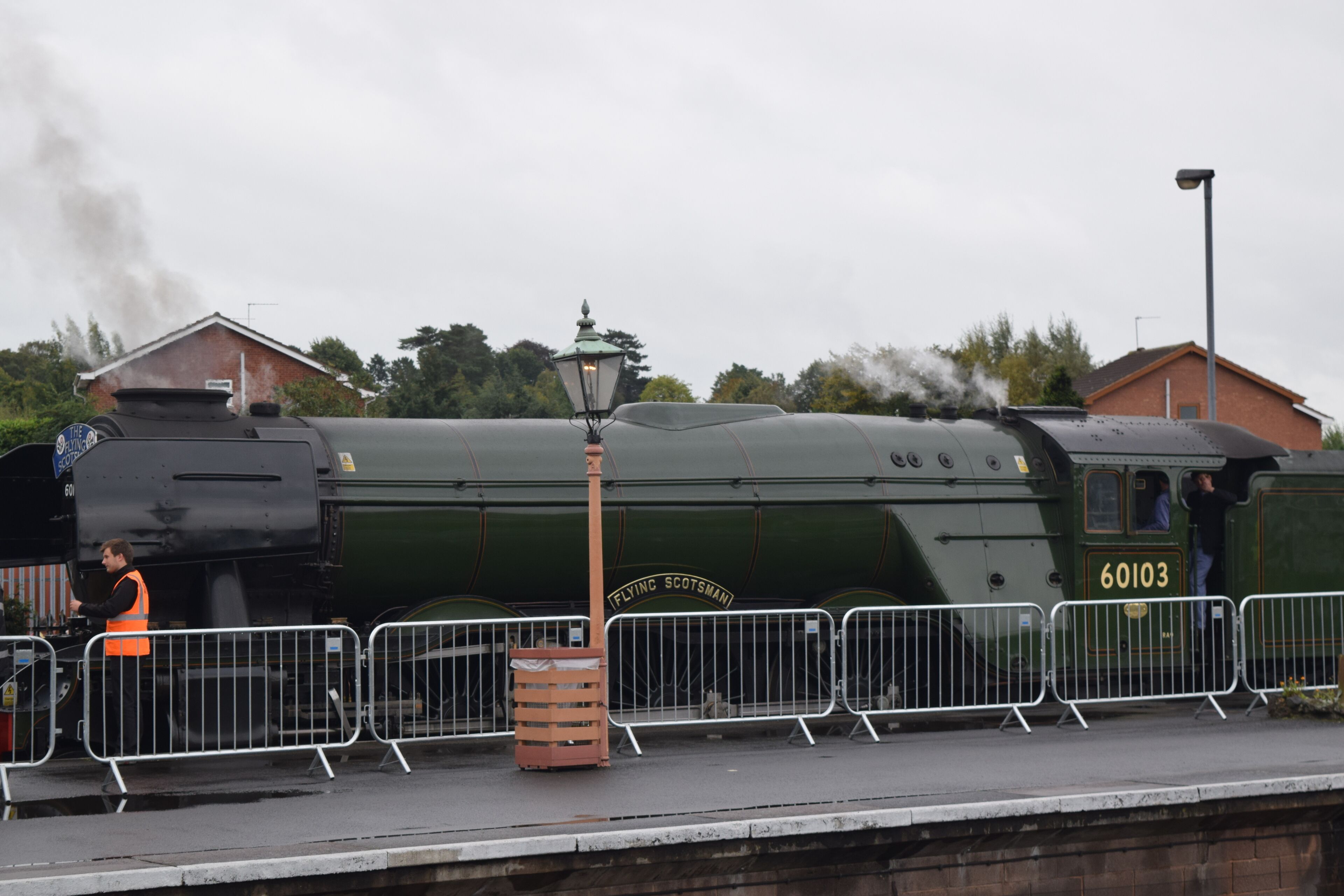 A vintage steam locomotive at a station with a platform, barriers, and a lamp post.