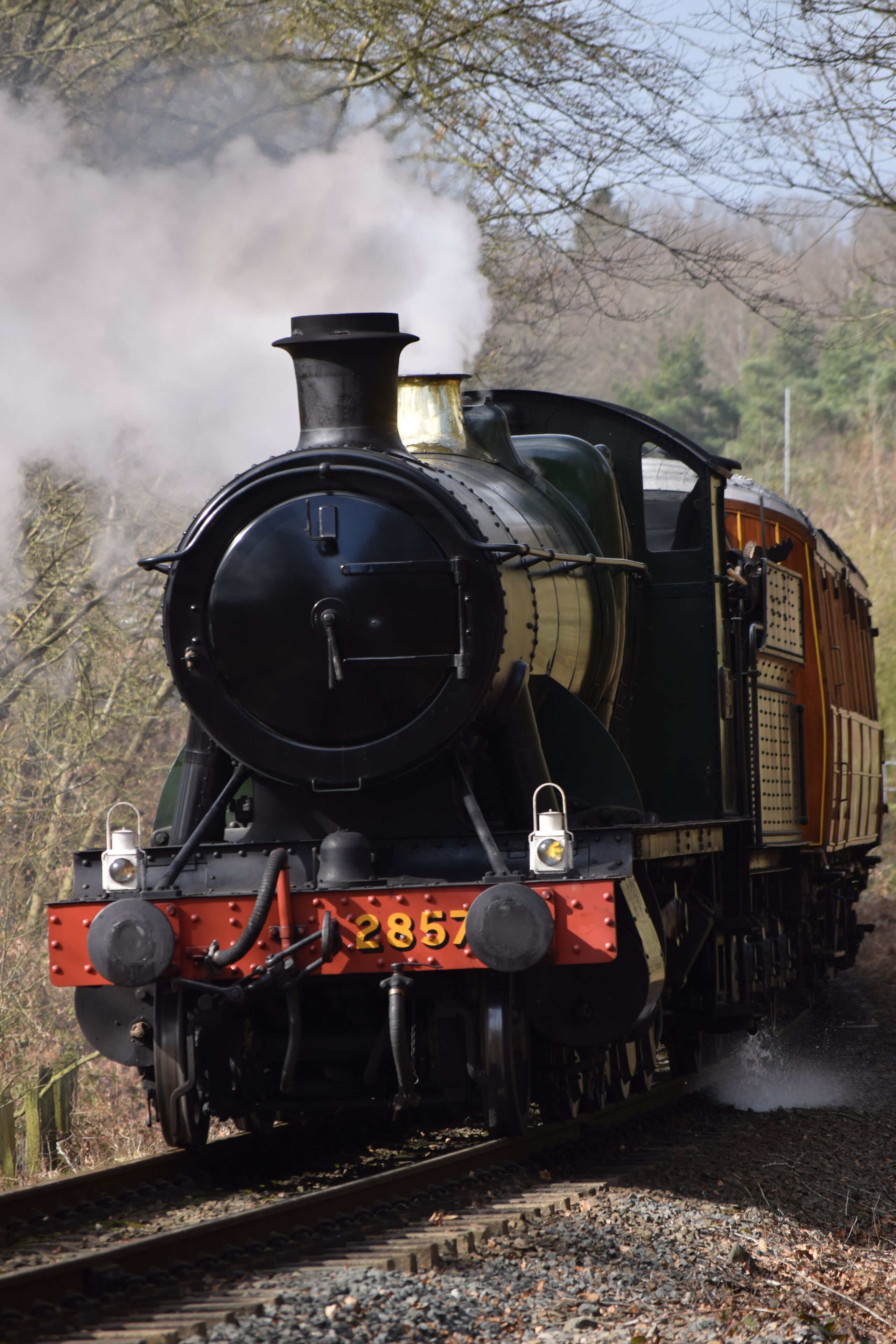 Una locomotora de vapor con el número 2857 en su parte frontal, circulando sobre una vía férrea rodeada de árboles.