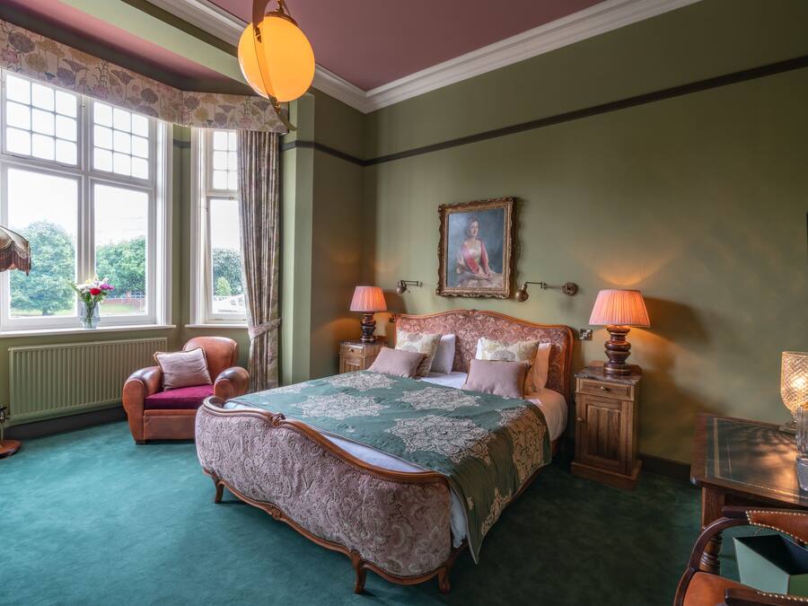 A traditional bedroom with a large bed, two bedside tables with lamps, a chair, and a window with curtains.