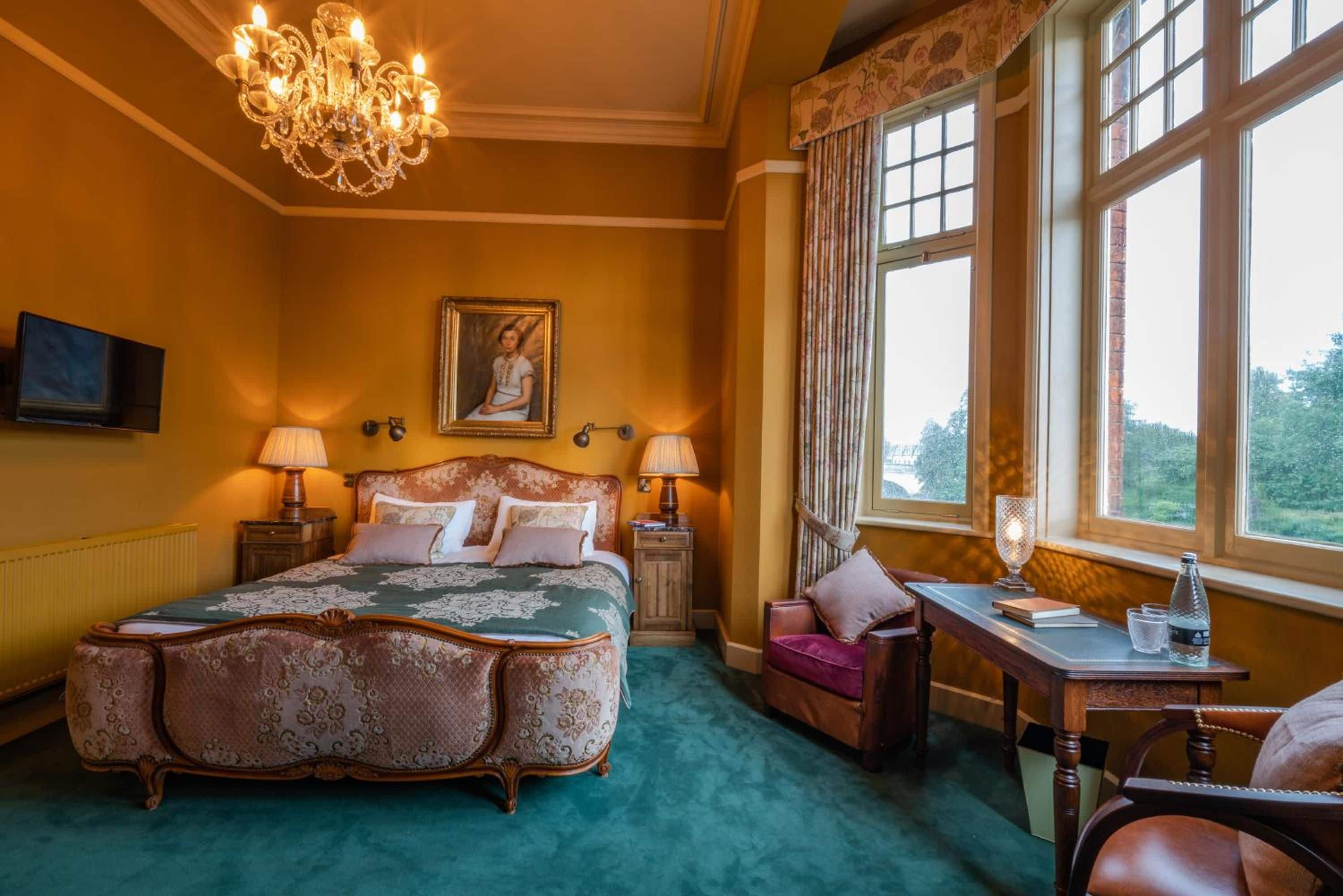 Ein Schlafzimmer mit einem großen Bett, zwei Nachttischen, einem Sessel, einem Schreibtisch, einem Fernseher und einem Fenster mit Vorhängen.