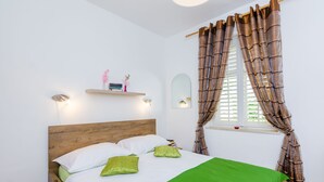 Een net opgemaakt bed met een houten hoofdbord, een groene sprei en twee kussens. Daarboven bevindt zich een plank met decoratieve objecten, en een raam met bruine gordijnen.