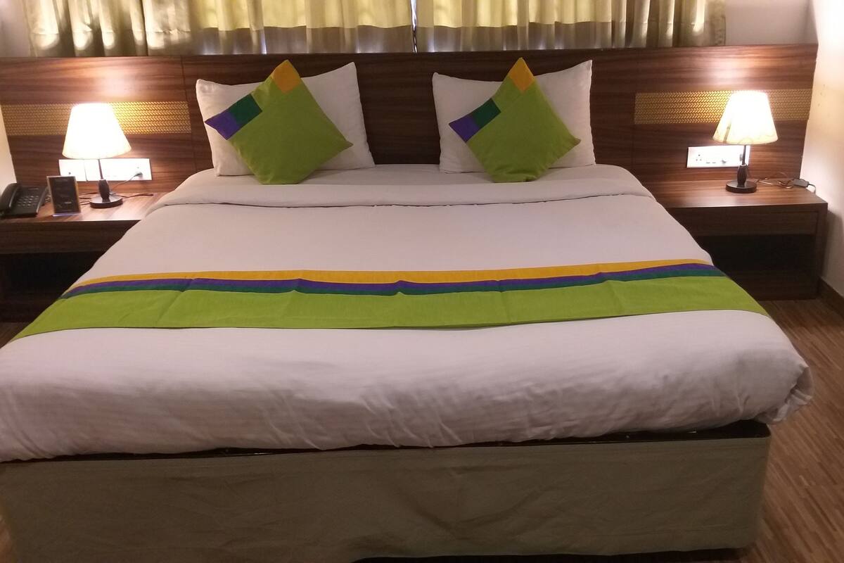 A neatly made bed with a white duvet, green and yellow striped blanket, and two green pillows. There are two bedside lamps on wooden nightstands.