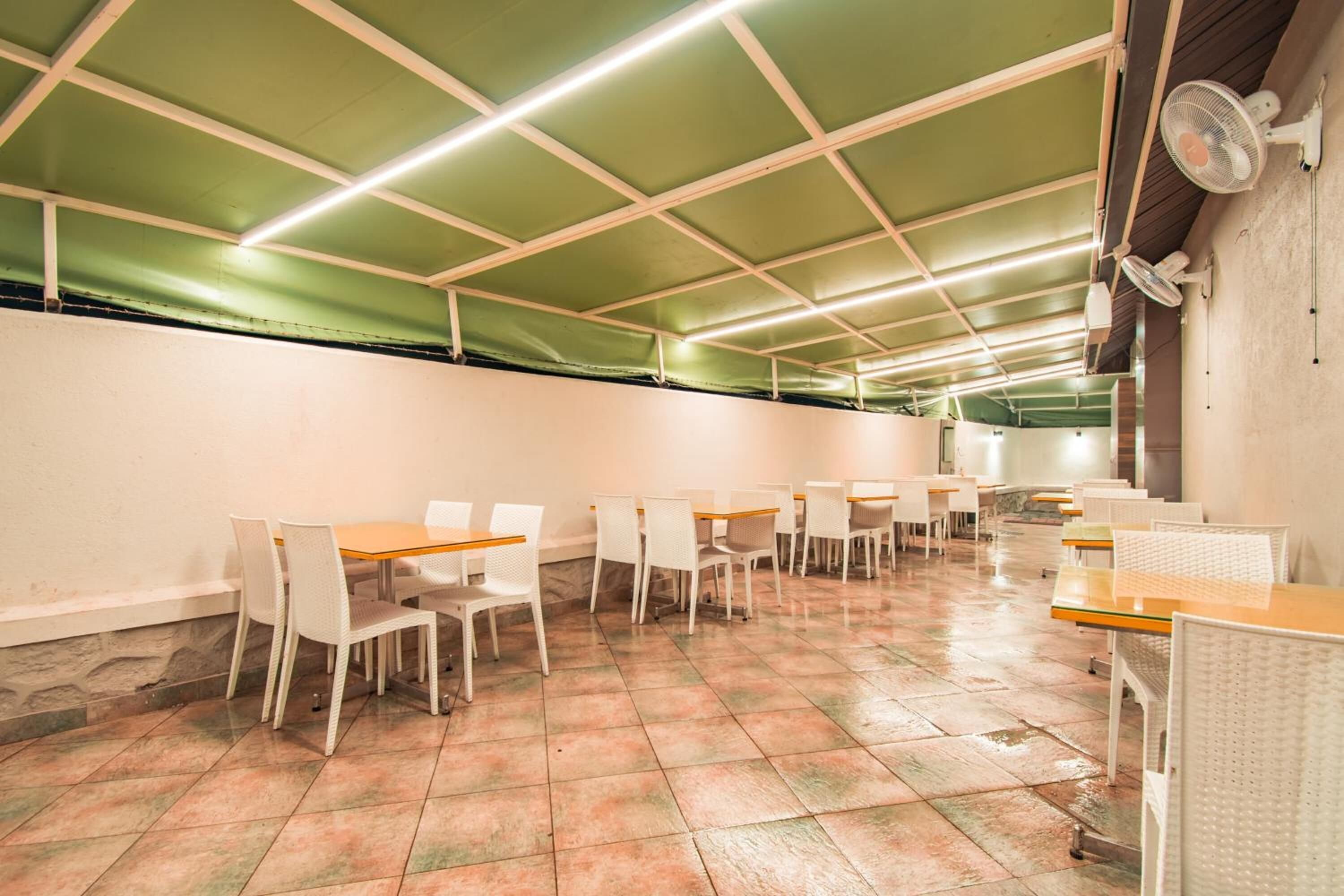 A spacious indoor area with multiple tables and chairs arranged in rows, a green ceiling with fluorescent lighting, and a fan mounted on the wall.