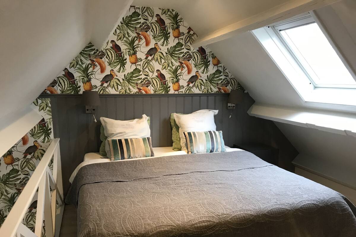 Une chambre avec un plafond en pente, un puits de lumière et un mur recouvert d’un papier peint représentant des oiseaux et des plantes tropicales.