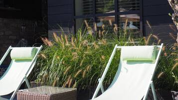 Duas cadeiras de descanso brancas com uma mesa de vime à frente, localizadas em frente a um edifício com grandes janelas e um jardim com grama alta.