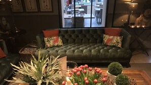 Un salon chaleureux avec un canapé vert, des coussins décoratifs, une table basse agrémentée de plantes et de bougies, et une vue sur la salle à manger à travers une porte vitrée.
