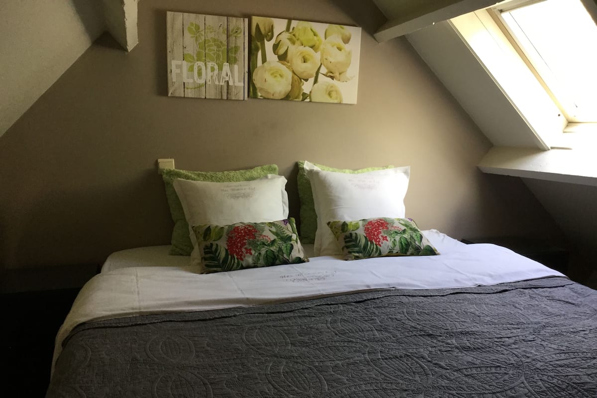 Une chambre avec un plafond en pente, un puits de lumière et un lit recouvert d’une courtepointe matelassée. On y trouve deux oreillers ornés de motifs floraux et un panneau mural sur lequel est inscrit le mot « FLORAL ».