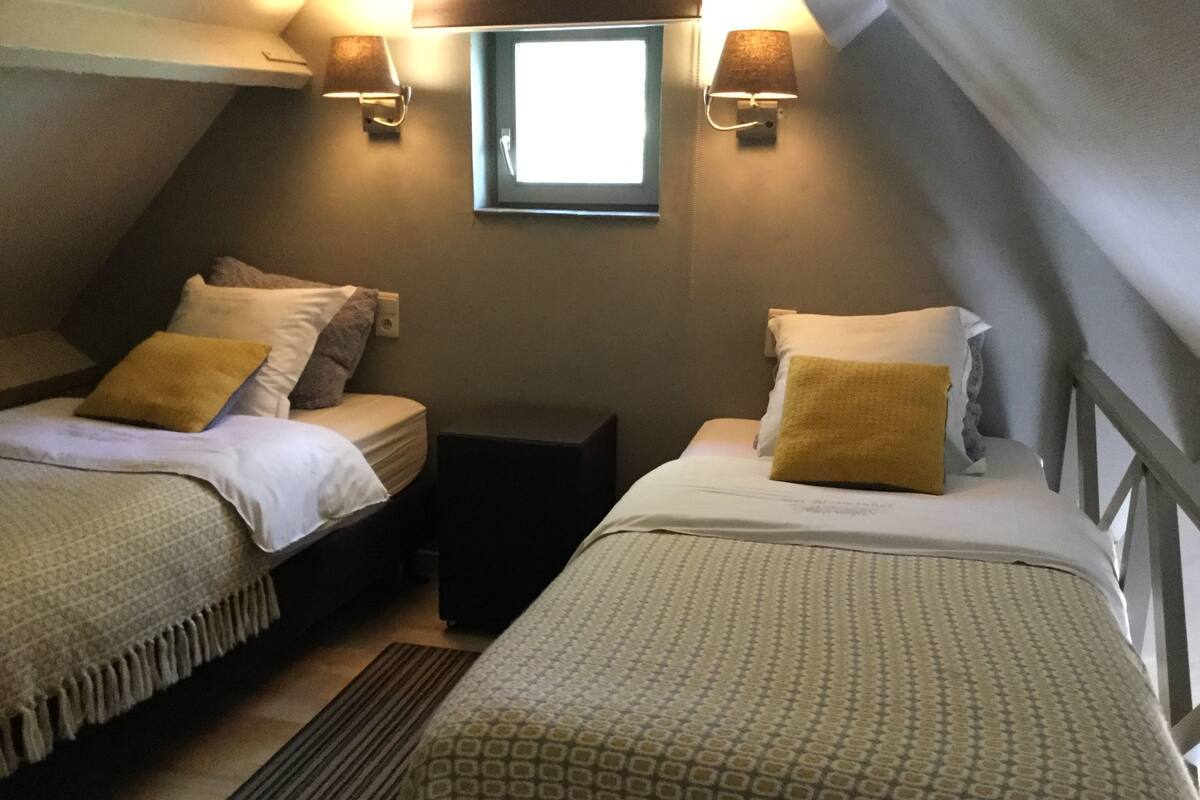 Une petite chambre accueillante avec deux lits, chacun muni d’un oreiller et d’une couverture. Une petite table se trouve entre les lits, et une fenêtre est située au-dessus de la table.