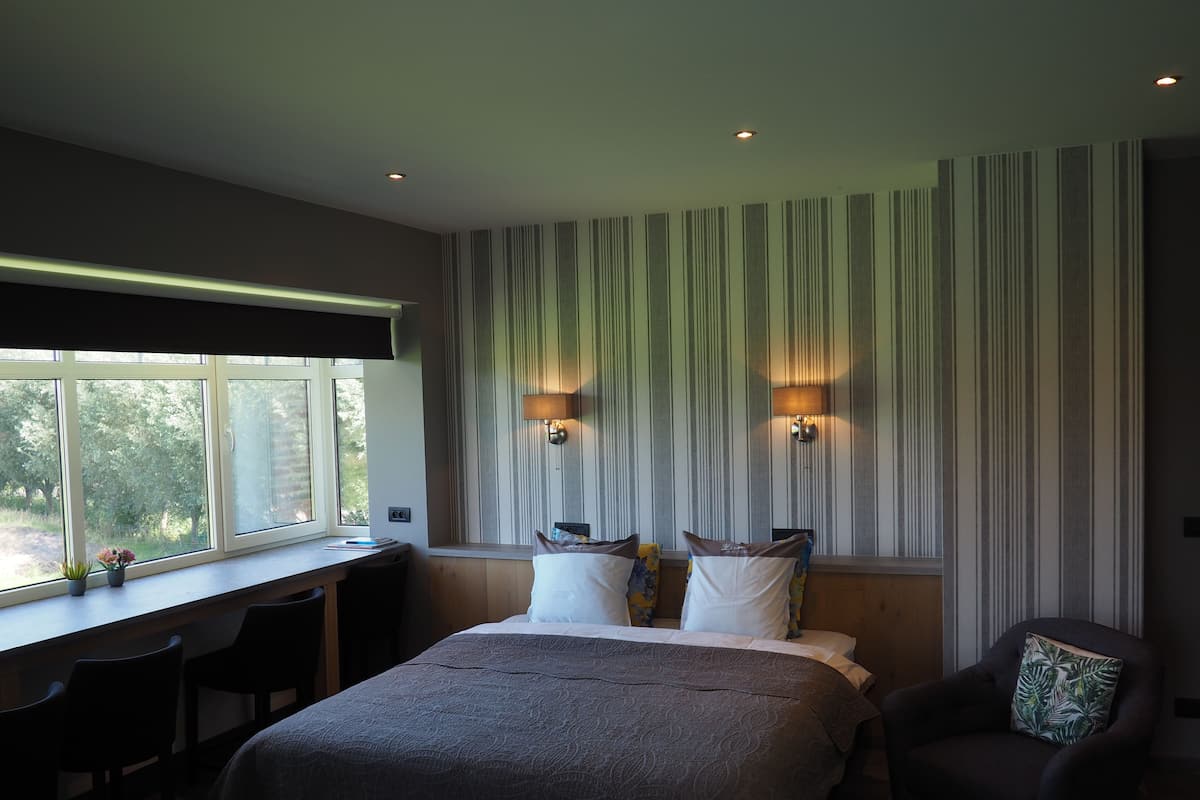 Une chambre à coucher avec un grand lit, un bureau, deux chaises, une fenêtre donnant sur un beau paysage et deux lampes fixées au mur.