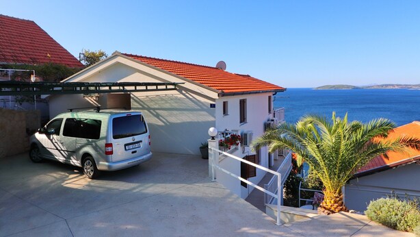 A white van parked in a driveway with a red-roofed building and a view of the sea.