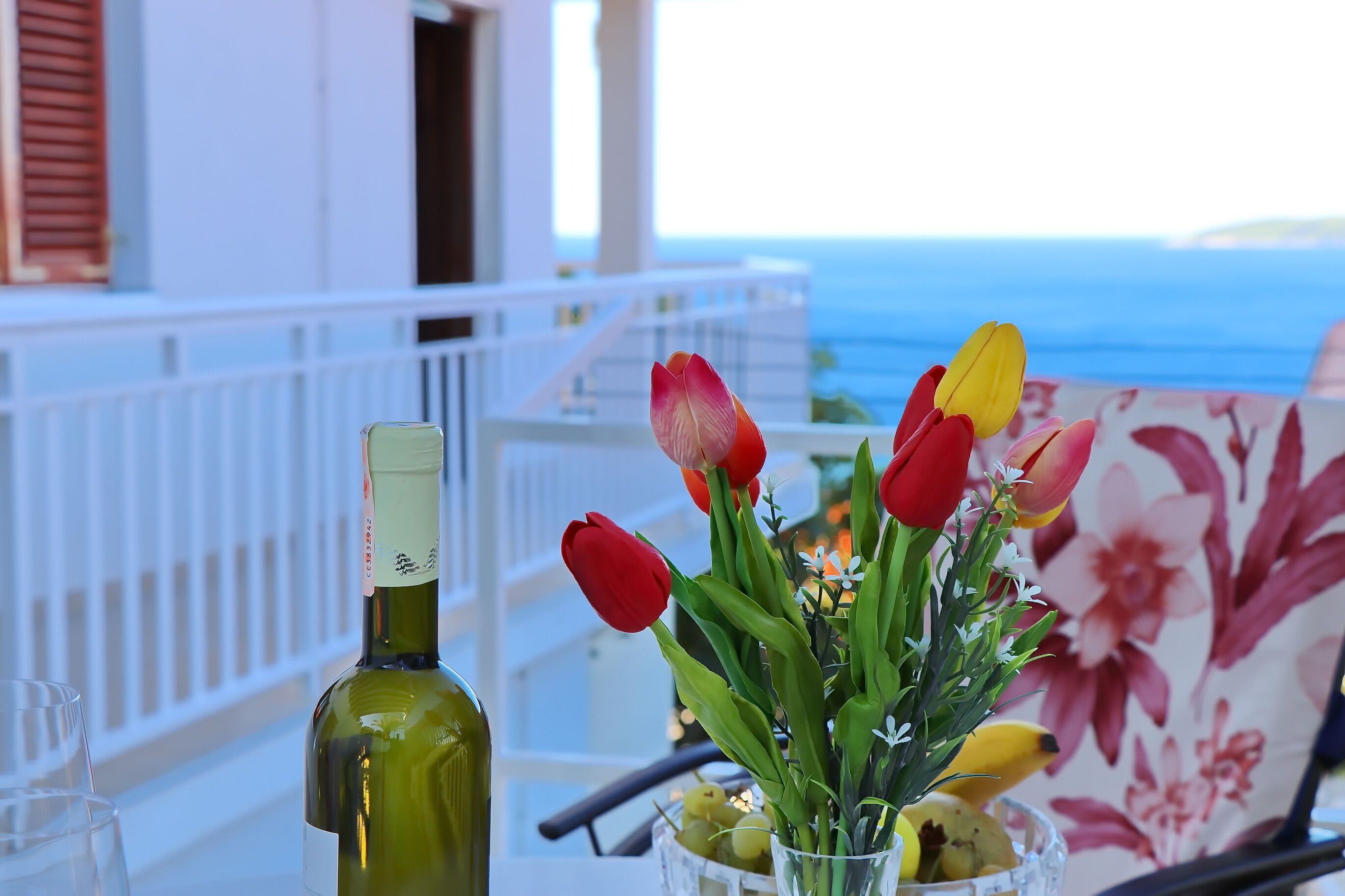 Een fles wijn, een glas, en een vaas met bloemen en fruit op een balkon met uitzicht op zee.