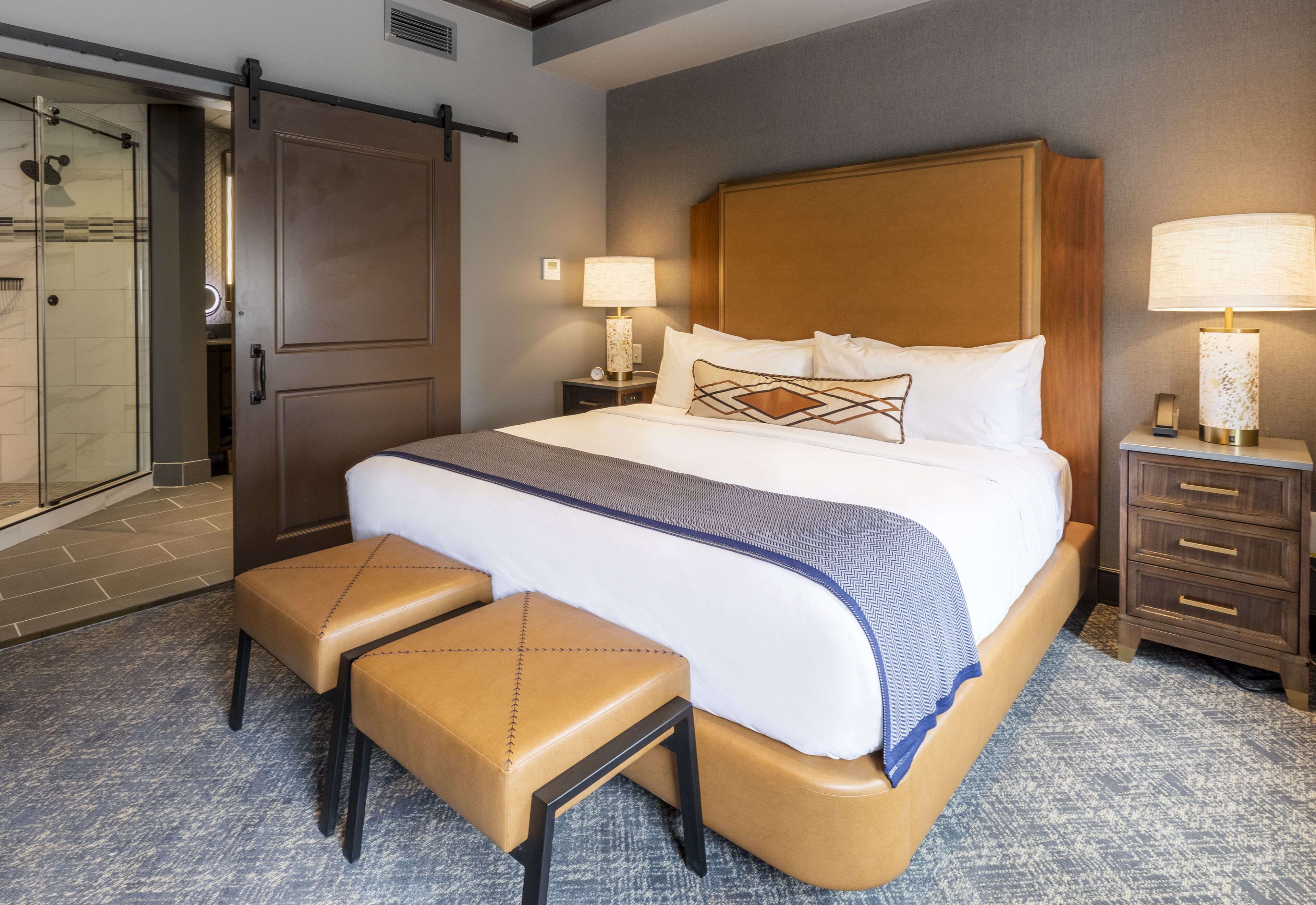 Habitación de hotel con una cama grande, un banco, una mesita de noche, una lámpara y una puerta corredera hacia la zona de la ducha.