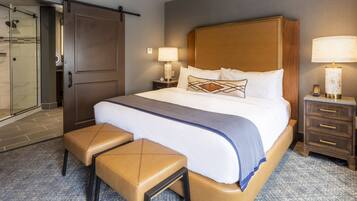Habitación de hotel con una cama grande, un banco, una mesita de noche, una lámpara y una puerta corredera hacia la zona de la ducha.
