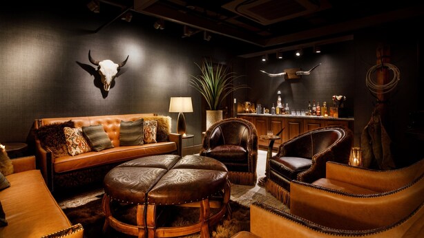 A dimly lit room with a leather sofa, a round wooden table, and leather armchairs. A bull skull is mounted on the wall, and there is a bar area in the background. - The Barfield, Autograph Collection (Amarillo)