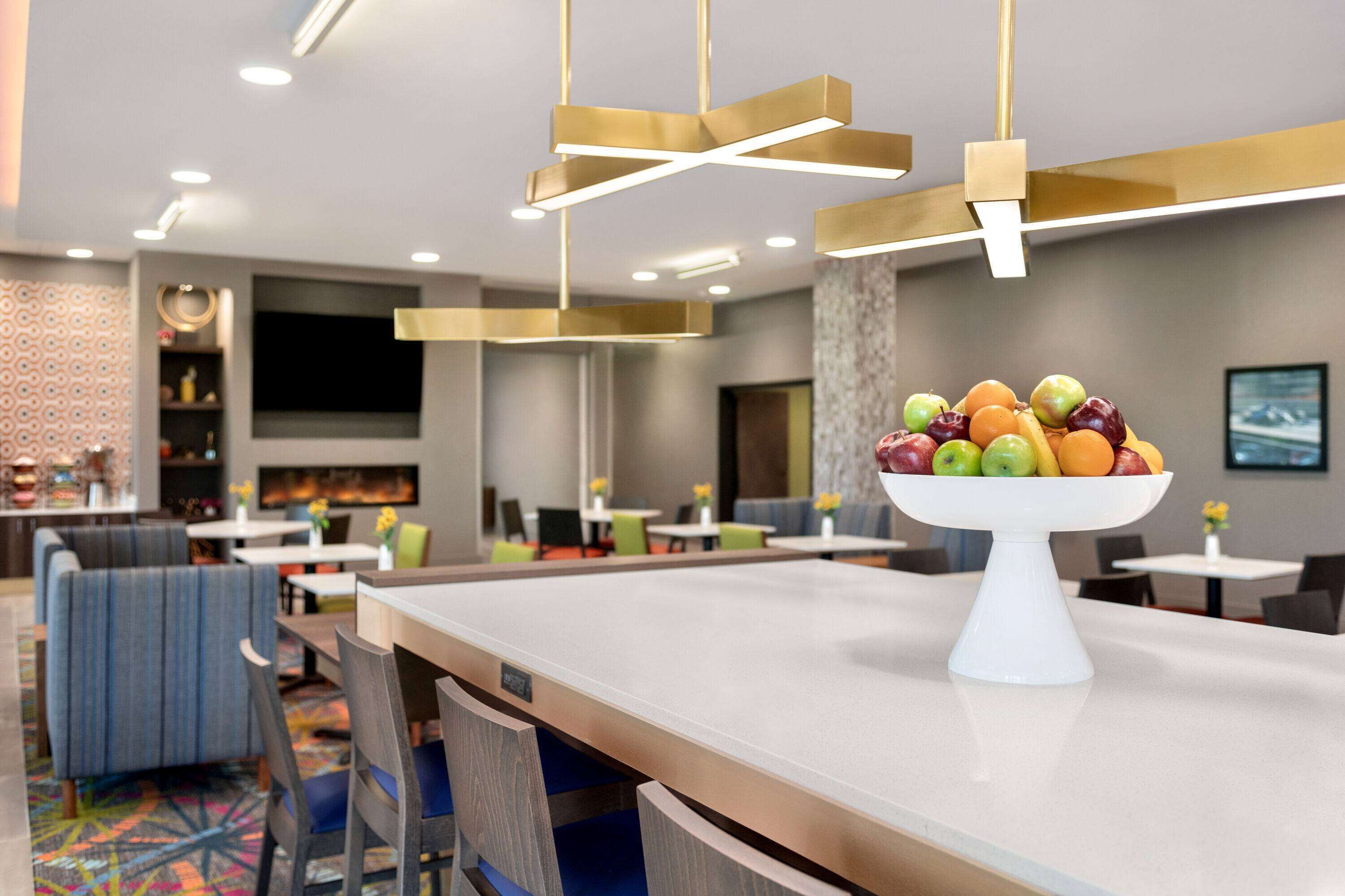 Um saguão de hotel moderno com área de bar, sofás e um recipiente com frutas no balcão.