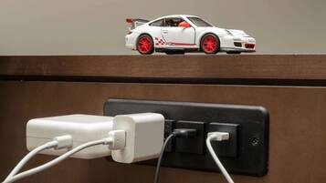 Une petite voiture jouet blanche, agrémentée de touches de rouge, est posée sur une étagère au-dessus d’une prise électrique murale dotée de deux fiches.
