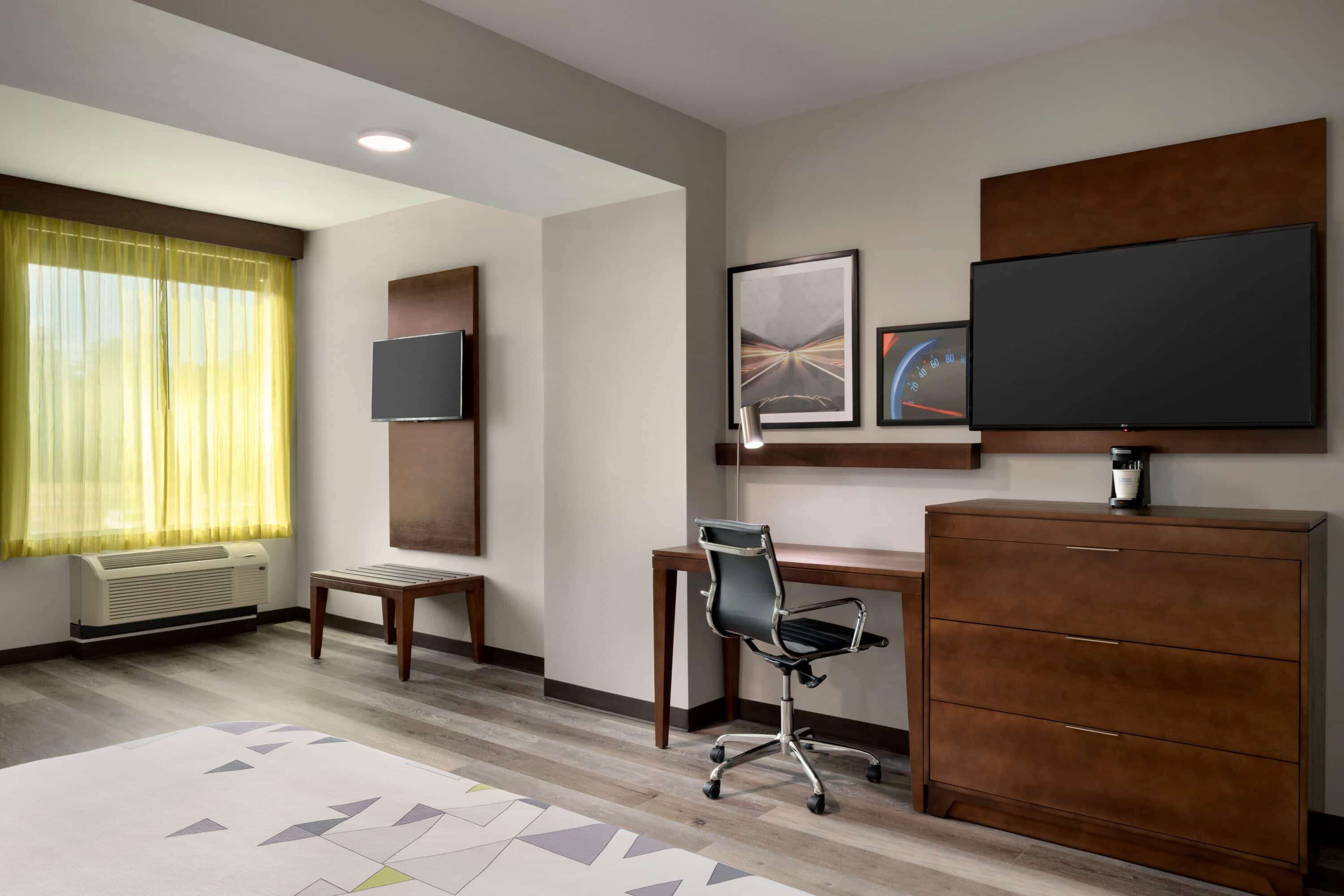 Habitación de hotel con escritorio, silla y un armario grande. Hay un televisor de pantalla plana en la pared y una ventana con cortinas.