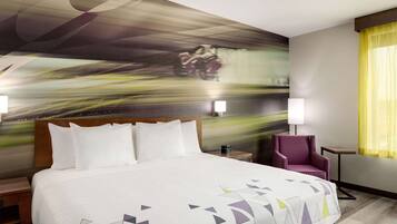 Ein Hotelzimmer mit einem großen Bett, einem Kopfteil, einem Nachttisch, einem Sessel, einer Lampe und einem Wandbild.