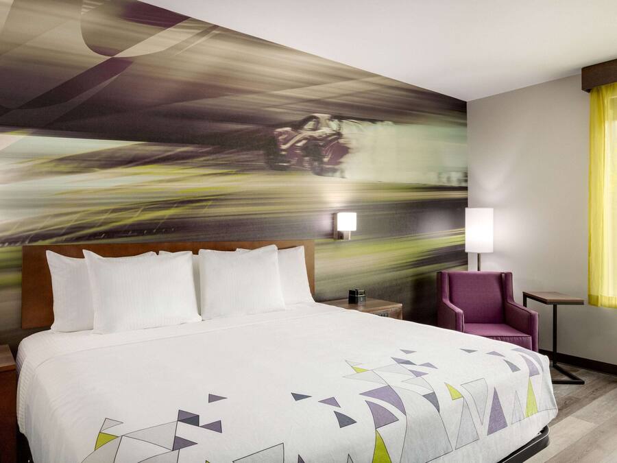 Ein Hotelzimmer mit einem großen Bett, einem Kopfteil, einem Nachttisch, einem Sessel, einer Lampe und einem Wandbild.