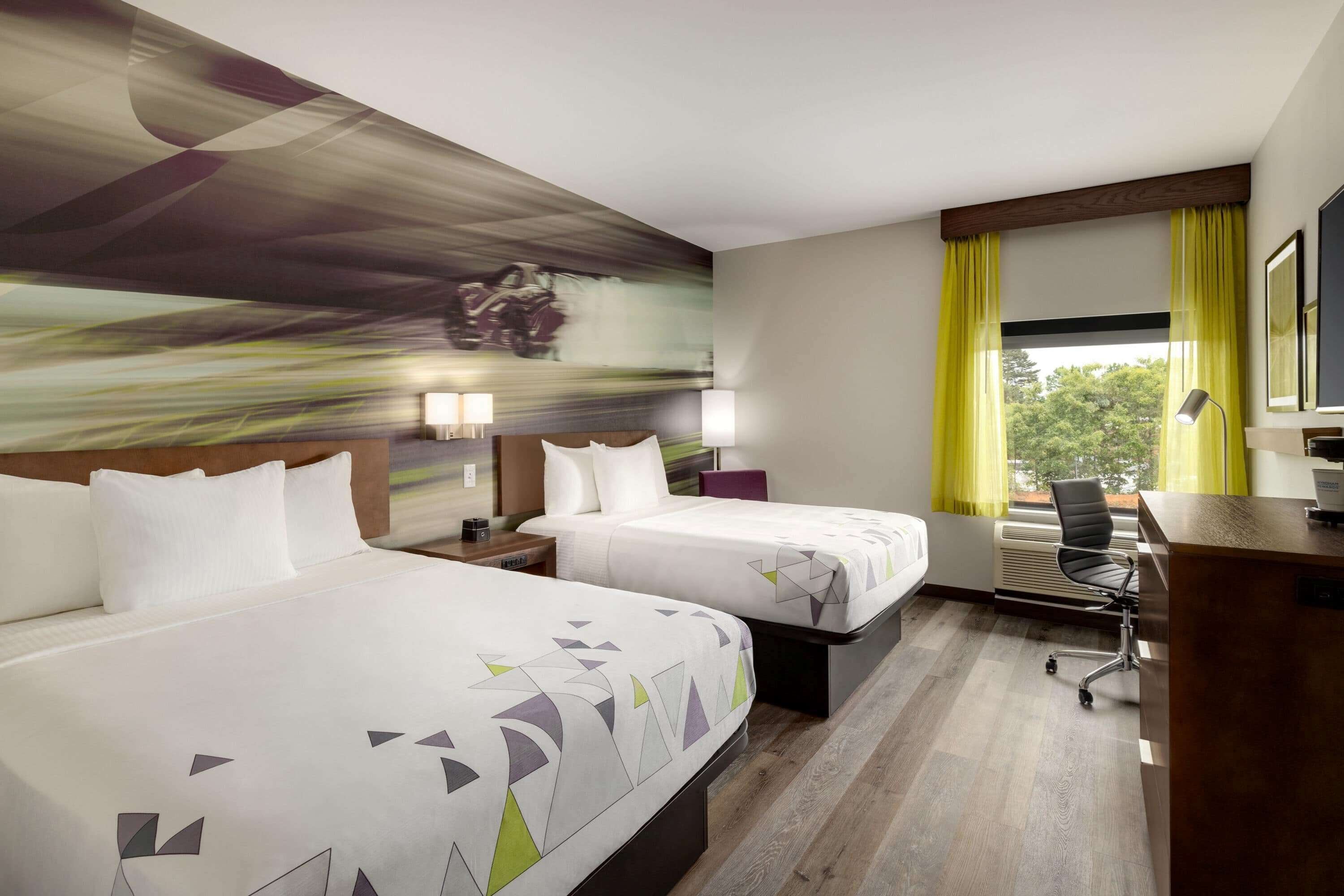 A hotel room with two beds, a desk, a chair, and a large window with curtains.