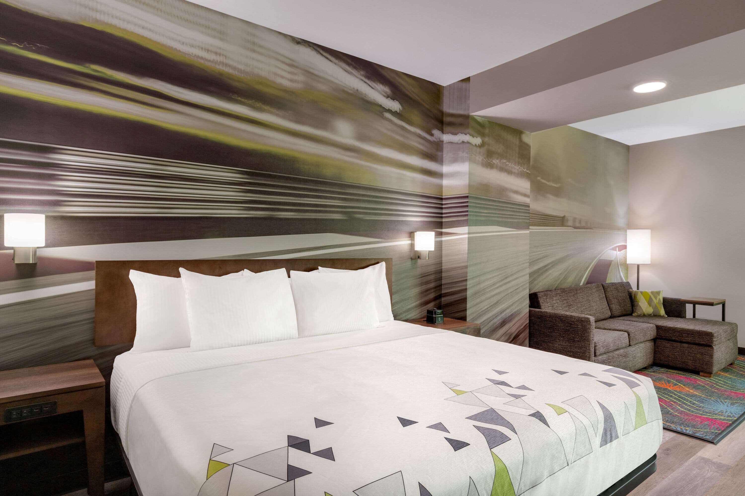Habitación de hotel con una cama grande, un sofá, una mesita de noche y una pared con un mural texturizado.