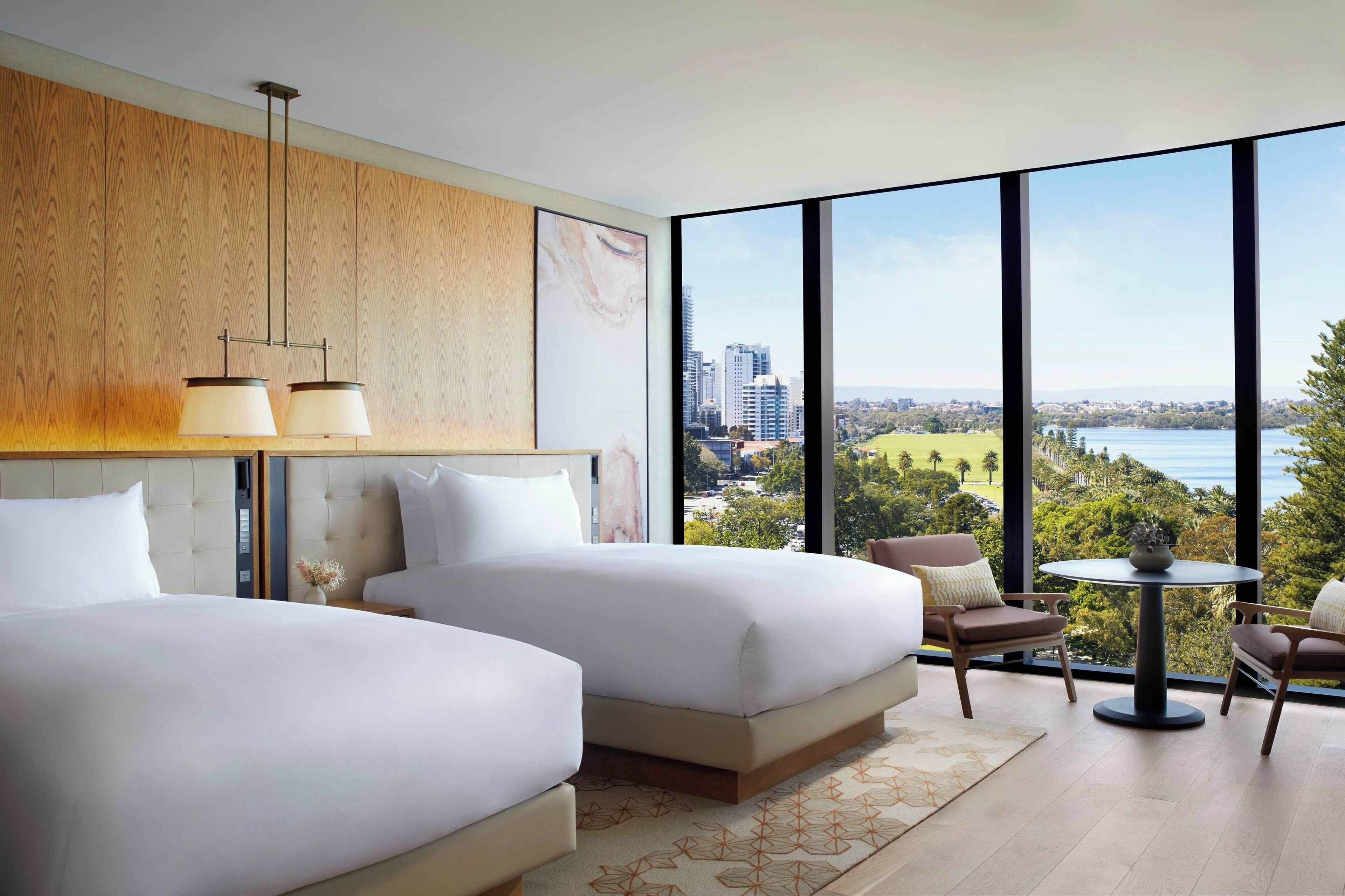 Een moderne hotelkamer met twee bedden, een eettafel en uitzicht op een stadsbeeld en water.