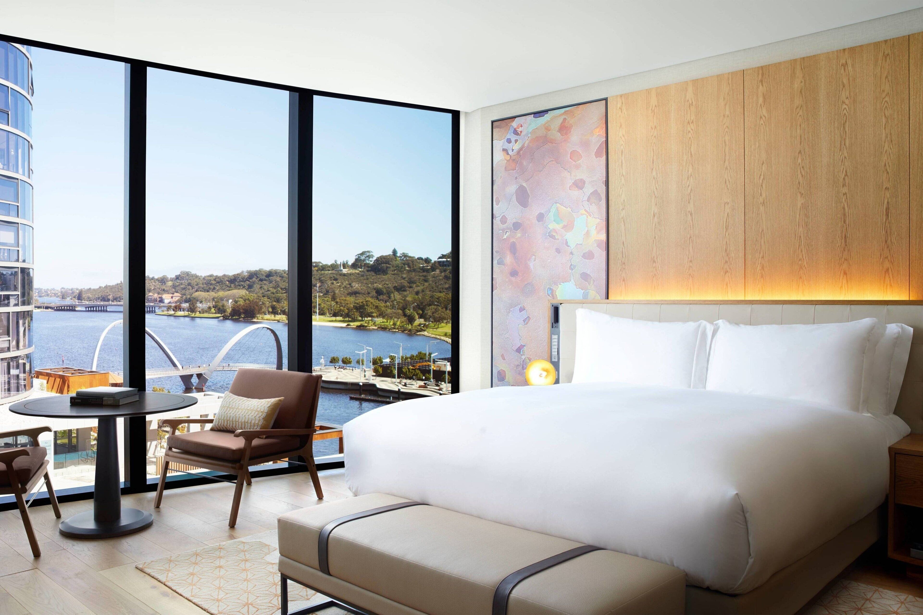 Una habitación de hotel moderna con una cama grande, vista a un cuerpo de agua, un panel de pared de madera y una zona de estar con una mesa y sillas.