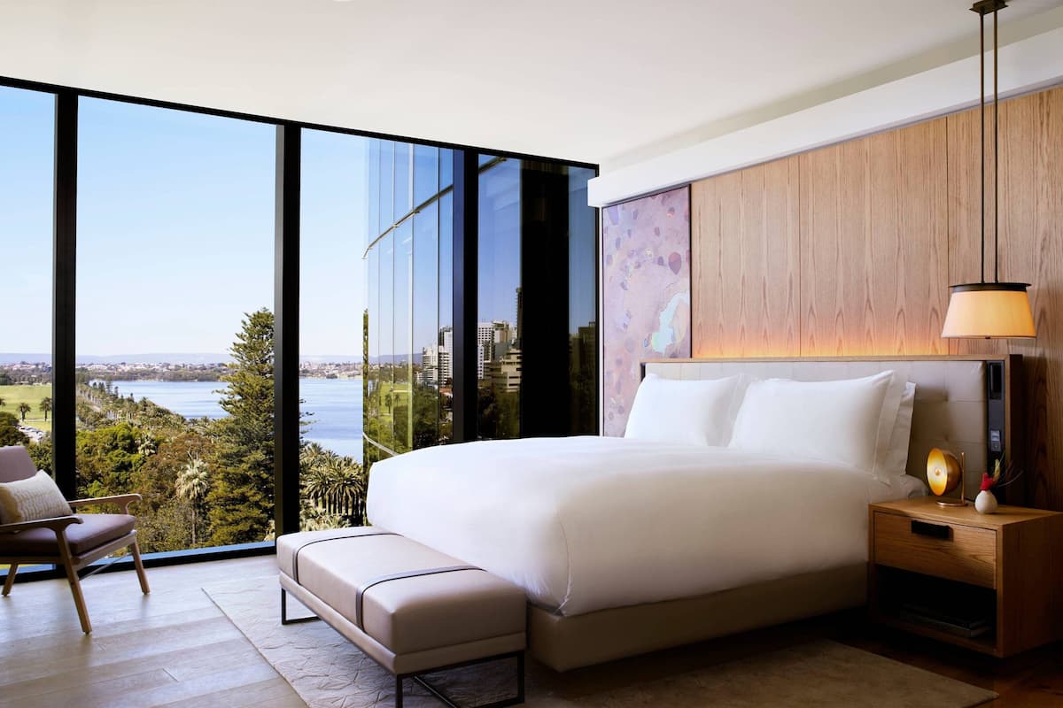 Un dormitorio moderno con una cama grande, una mesita de noche de madera, un banco y vistas a un paisaje urbano y al agua a través de ventanales.