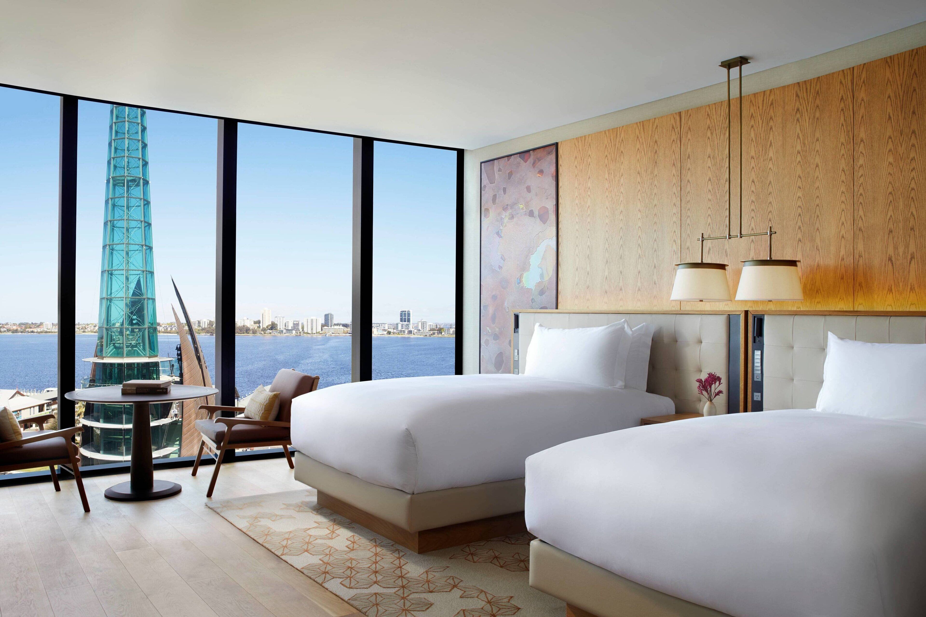 A modern hotel room with two beds, a large window offering a city view, a dining table, and a view of a tall building.