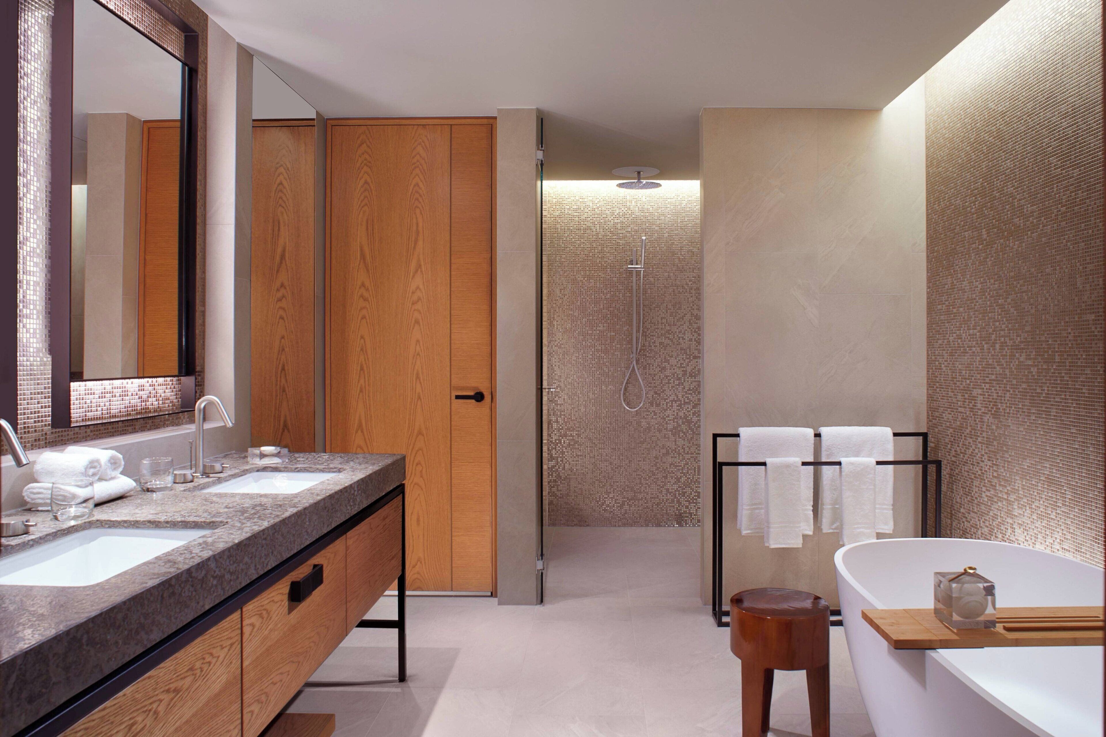 Un baño moderno con dos lavamanos, un espejo grande, una puerta de madera, un box de ducha con paredes de vidrio y una bañera con un taburete de madera.