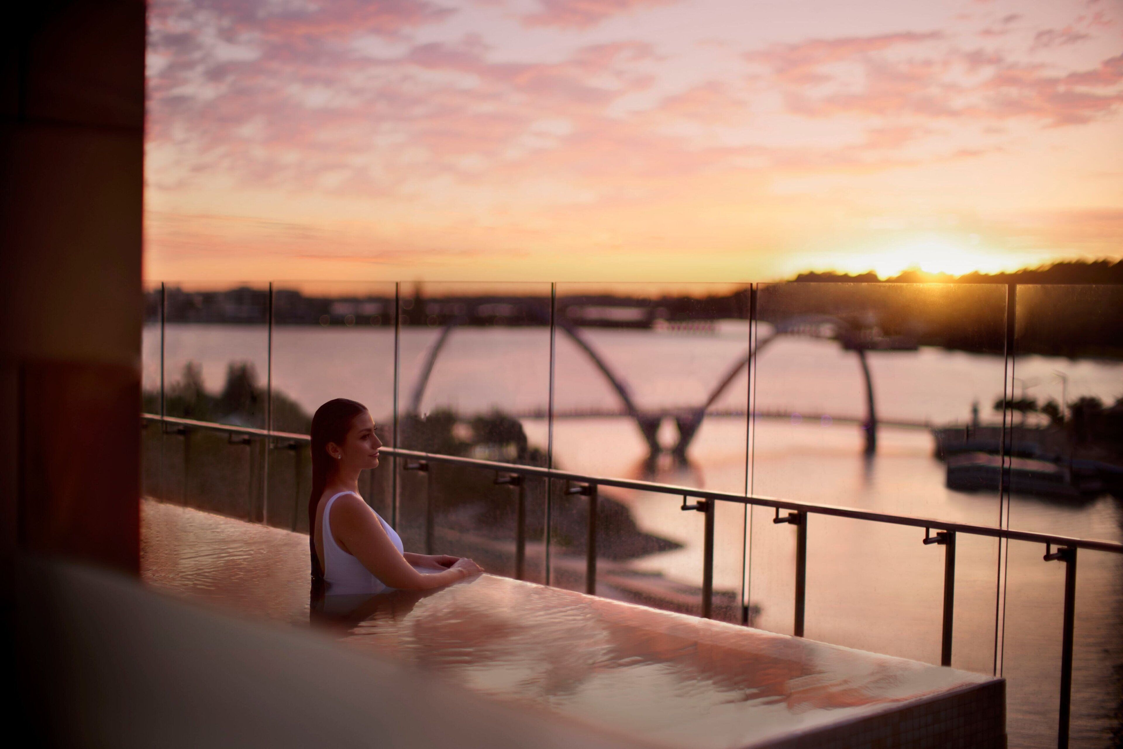 A person sitting by a railing overlooking a sunset over a river with a bridge in the distance.