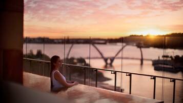 A person sitting by a railing overlooking a sunset over a river with a bridge in the distance.