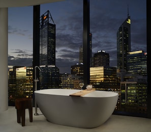 A modern bathroom with a freestanding bathtub, a wooden stool, and a large window offering a cityscape view at dusk.