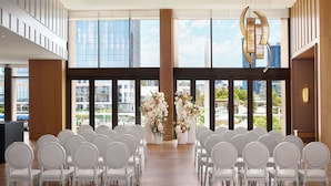 A modern indoor venue with rows of white chairs arranged for an event, large windows offering a city view, and decorative floral arrangements.