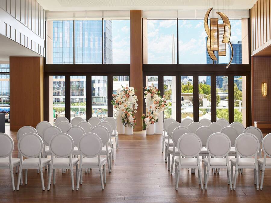 A modern indoor venue with rows of white chairs arranged for an event, large windows offering a city view, and decorative floral arrangements.