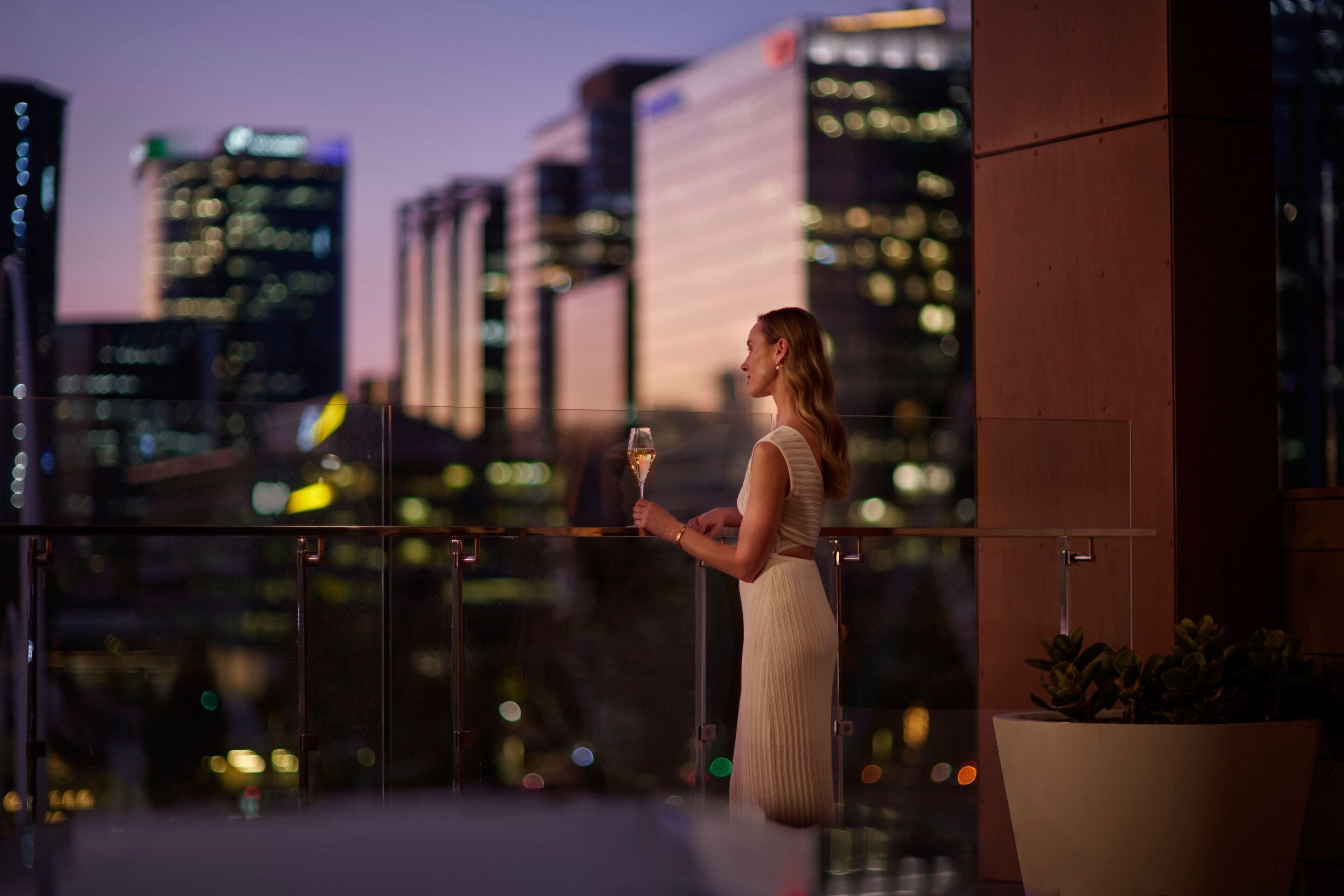 Una mujer con un vestido blanco de pie en un balcón con vistas a la ciudad.